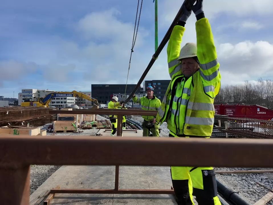 Arbeidere i verneutstyr løfter på plass jernstang som henger fra kran